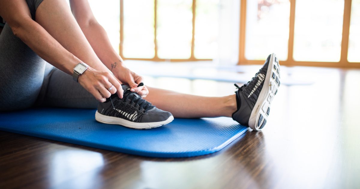 A woman sitting on a blue exercise mat tying her running shoes in a sunny studio