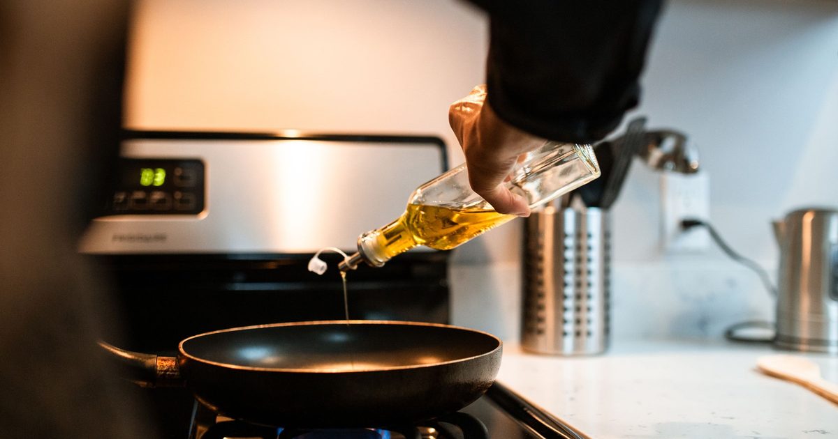Olive oil being poured from a glass bottle into a hot frypan on a gas stove