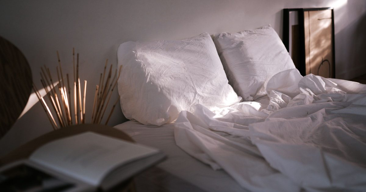 A neatly made white bed with an open book and a reed diffuser in soft evening light